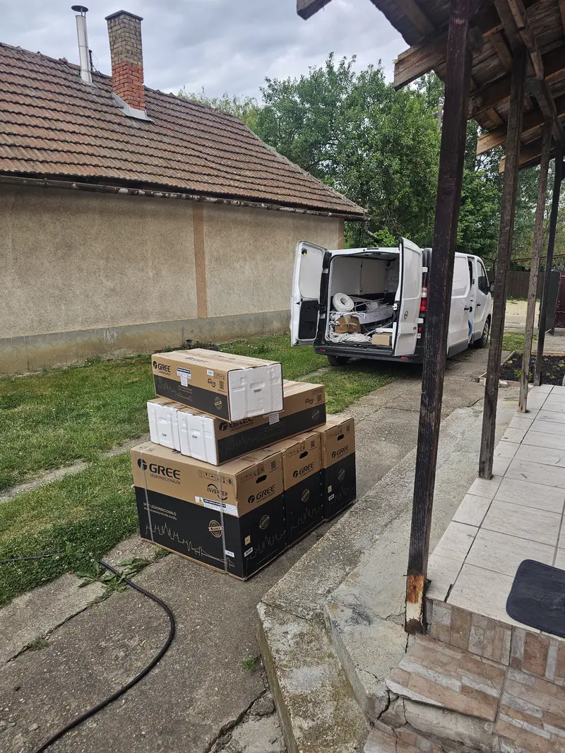 Gree klímák dobozban egy udvaron, mögöttük a szerelők nyitott furgonja